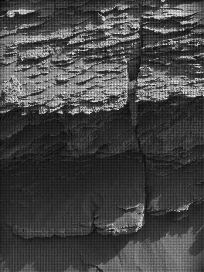 A grayscale photo from the Martian surface shows a rocky outcrop rising from sandy ground, all of it medium gray. The rock feature is composed of thin, rough-edged layers, stacked on top of each other, rising from the ground to the top of the frame. The layer at bottom sticks out farther, toward the viewer, and is thicker than the rest and covered in a layer of sand.