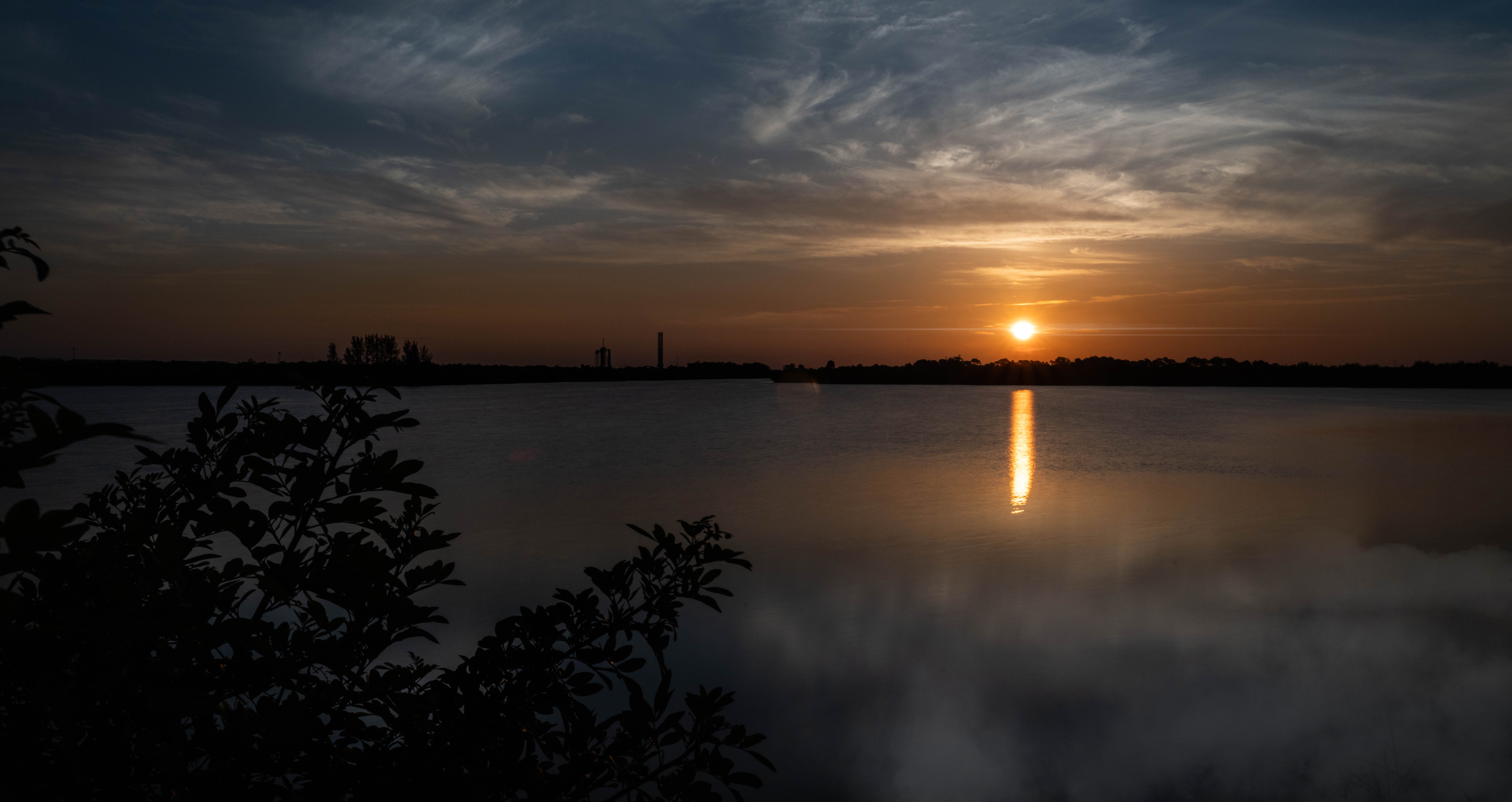 In the distance, the Sun rises, casting an orange glow over the surrounding sky. It is also reflected in the water below it, where it looks like a column of orange light. The outlines of several structures are visible in the middle ground. At the top of the photo are white clouds that look like streaks of paint. At the bottom of the photo, those clouds are partially reflected in the water (at right) and some foliage peeks in (at left).