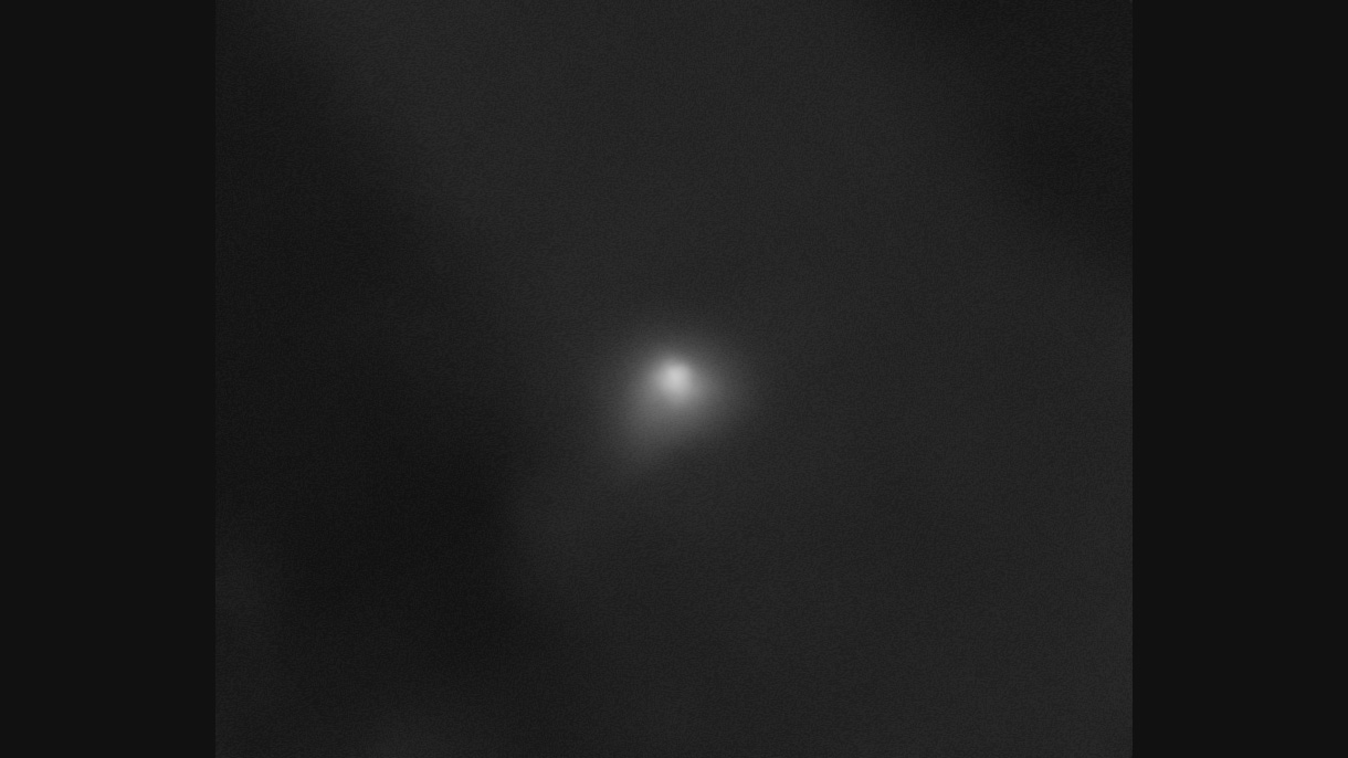On a largely black background, interstellar comet 3I/ATLAS appears as a white smudge with a semicircular shape at its core. A transparent white cloud extends slightly from the comet toward the bottom left of the image and, to a lesser extent, toward the right side of the image.