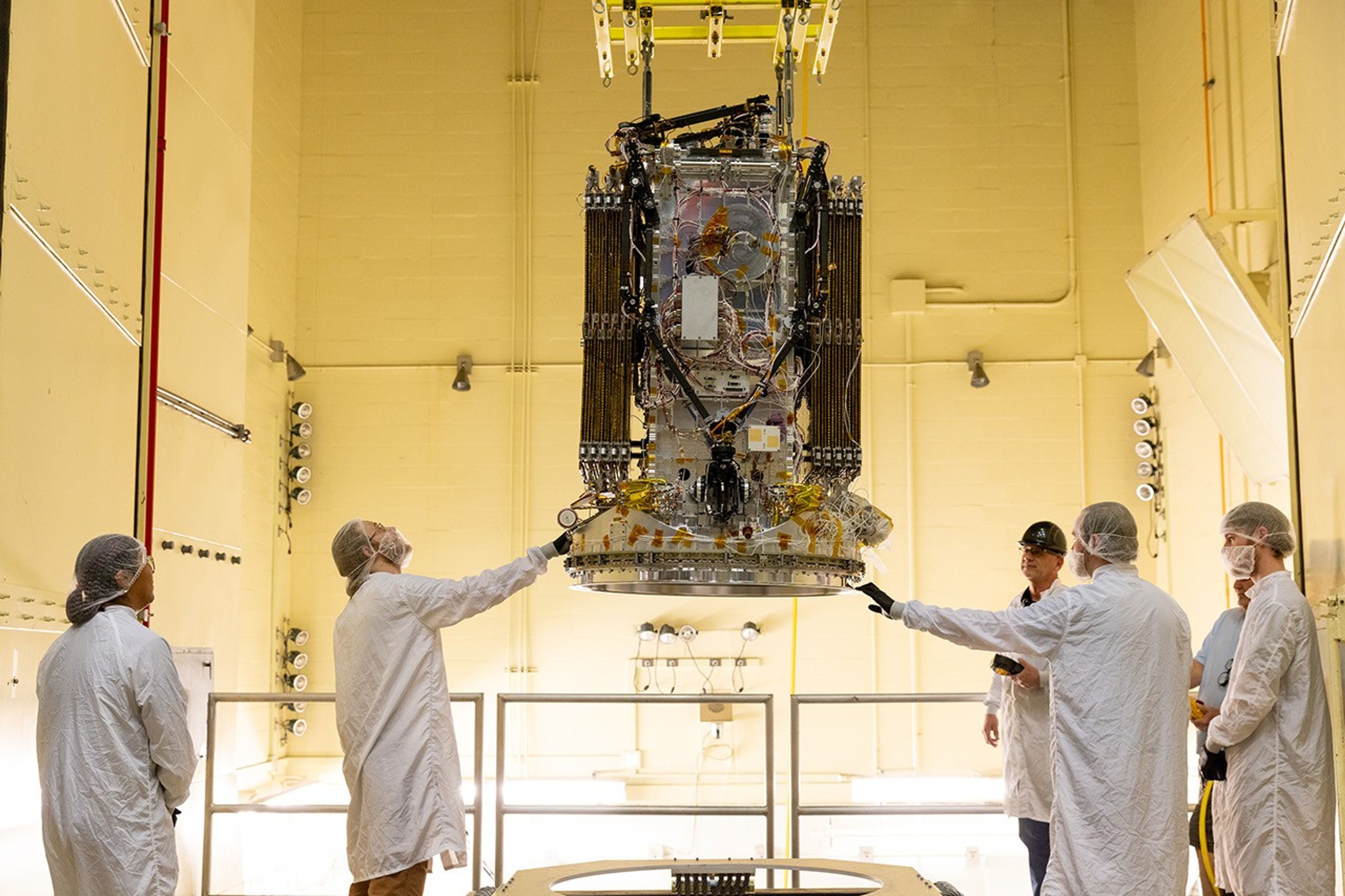 Staff in white lab coats observe a black and gray spacecraft in a room with warm, yellow light.